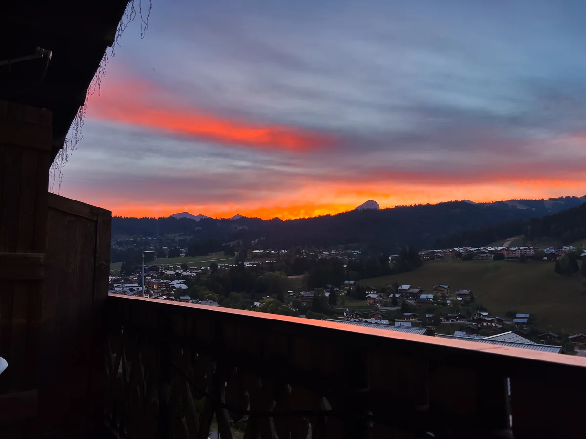 Lever de soleil orange et rose sur le village et les collines boisées, vu d'un balcon de chalet en bois