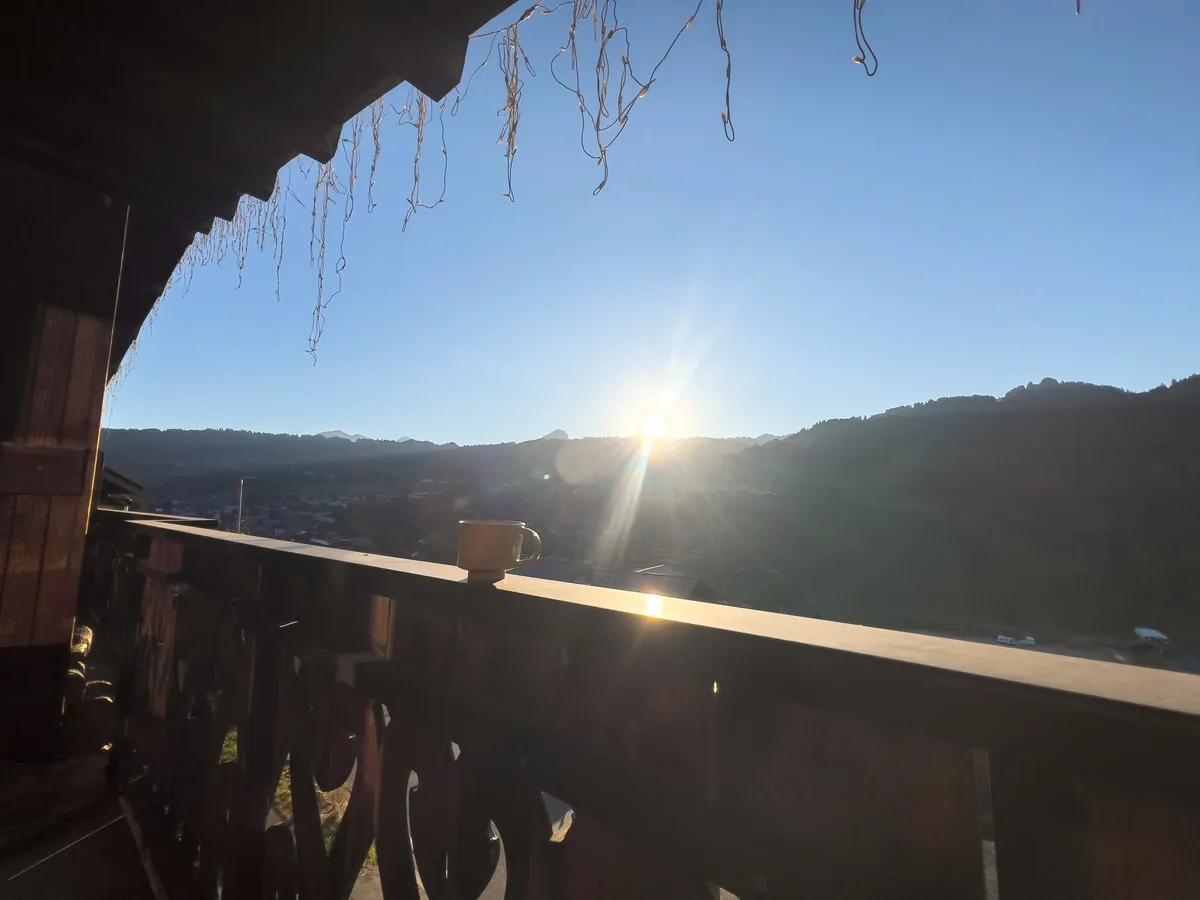 Le soleil se lève sur les montagnes, vu depuis un balcon de chalet en bois avec une tasse posée sur la rambarde.